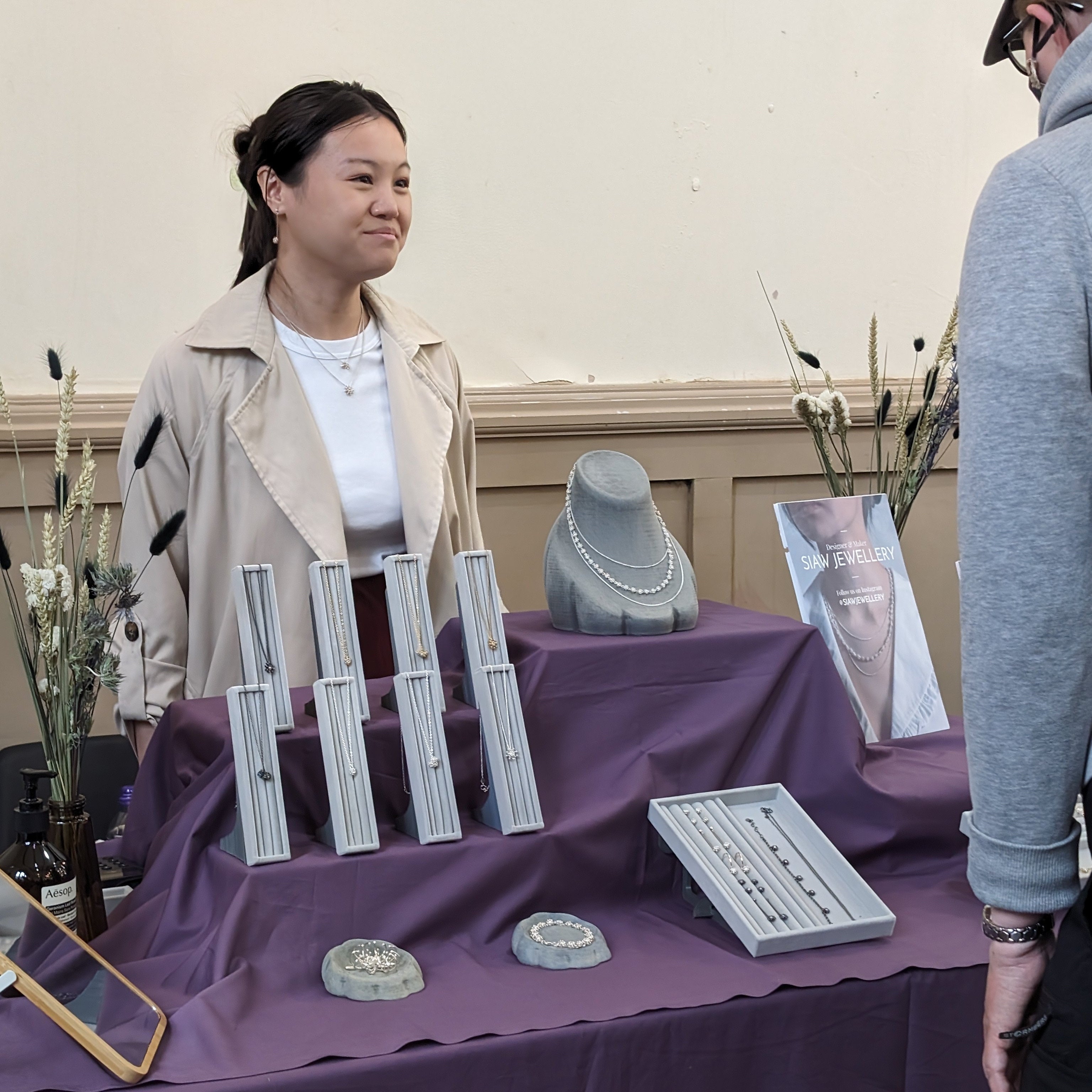Woman standing behind a table with jewelry displays, interacting with another person.